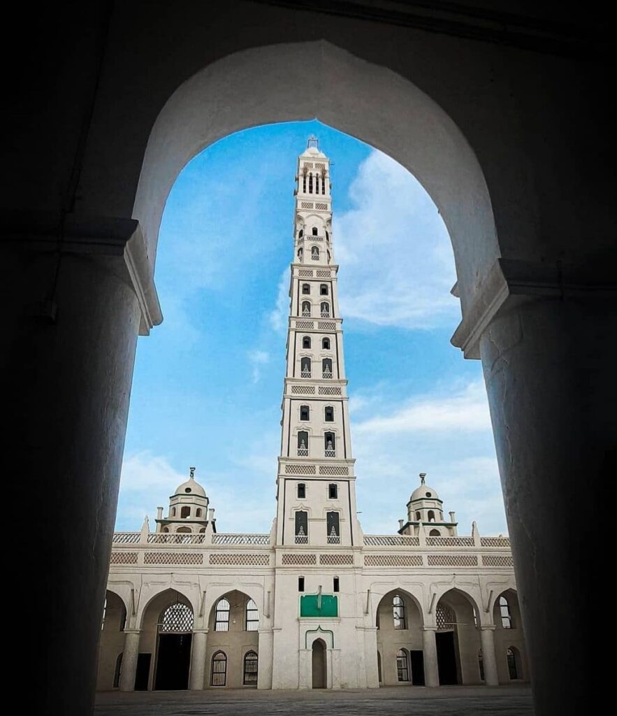  A front-facing, full-view shot of the Al-Mihdhar Mosque in Tarim, highlighting its majestic white facade and iconic tall minaret against a clear blue sky.